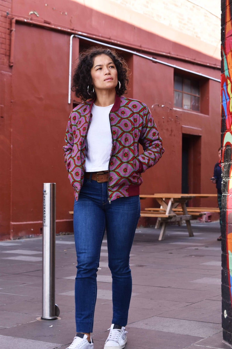 May include: A woman wearing a patterned bomber jacket with a white t-shirt and blue jeans. The jacket features a pink and red geometric design. She is also wearing a brown belt and white sneakers. The background includes a red brick wall and a graffiti-covered wall.