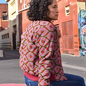 May include: A pink bomber jacket with a geometric pattern in red, green, and yellow. The jacket has a burgundy collar, cuffs, and waistband. The person is wearing blue jeans.