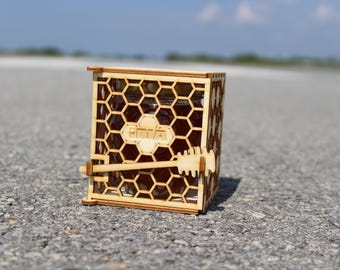 May include: A wooden honey jar with a honeycomb design and a wooden honey dipper. The jar is labelled "PAVA".