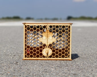 May include: A wooden box with a honeycomb design, containing two glass jars of honey. The box has a label that reads "PRVA KUĆA MEDA ZUPANIJA".