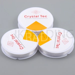 May include: Three spools of clear elastic beading cord. Each spool is on a white circular card with the words "Crystal Tec" in red and a yellow triangle design. The spools are stacked on a grey surface.