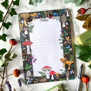 May include: A notepad with a black background and a colorful border featuring various mushrooms, berries, and foliage. The word "Notes" is written at the top, and the pages are lined. A pen and book are also visible.