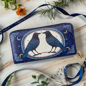 May include: A blue wallet with a design of two black ravens facing each other, with blue and white details. The ravens are set against a white background within an oval frame, with moons and stars. The wallet has a gold zip.