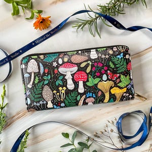 May include: Black wallet with a colorful mushroom and botanical design. The wallet features a variety of mushrooms, berries, and leaves. The zipper is gold-colored. The text "Mystical Sky Studio" is on a blue ribbon.