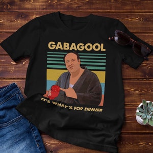 以下が含まれることがあります： 「GABAGOOL」と「IT'S WHAT'S FOR DINNER」の文字と、赤いカップを持った男性のレトロなグラフィックがプリントされた黒いTシャツ。木製の表面にジーンズ、サングラス、小さな植物と一緒に置かれています。
