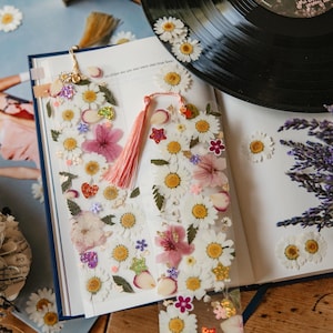 May include: Two clear rectangular bookmarks featuring embedded pressed white daisy flowers, pink petals, and small colorful glitter shapes. Each bookmark is finished with a soft pink tassel attached to the top, resting on an open book page.