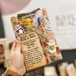 May include: A brown paper-wrapped book with a panda sticker, a pen, and a chocolate bar. The book is labeled "Blind Date With A Book" and has a description for a middle grade fantasy book.