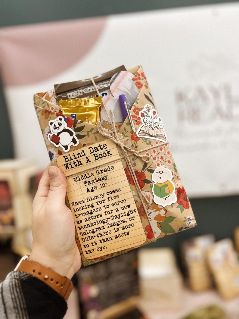 May include: A brown paper-wrapped book with a panda sticker, a pen, and a chocolate bar. The book is labeled "Blind Date With A Book" and has a description for a middle grade fantasy book.