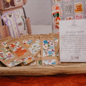 May include: A collection of rectangular paper bookmarks with floral patterns displayed in a wooden bowl. A white sign stands in the bowl with the text Reads & Seeds, describing the bookmarks as plantable wildflower seed paper priced at $18.