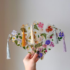May include: Set of five pressed flower bookmarks with different floral designs and colors. Each bookmark has a tassel attached to the top.