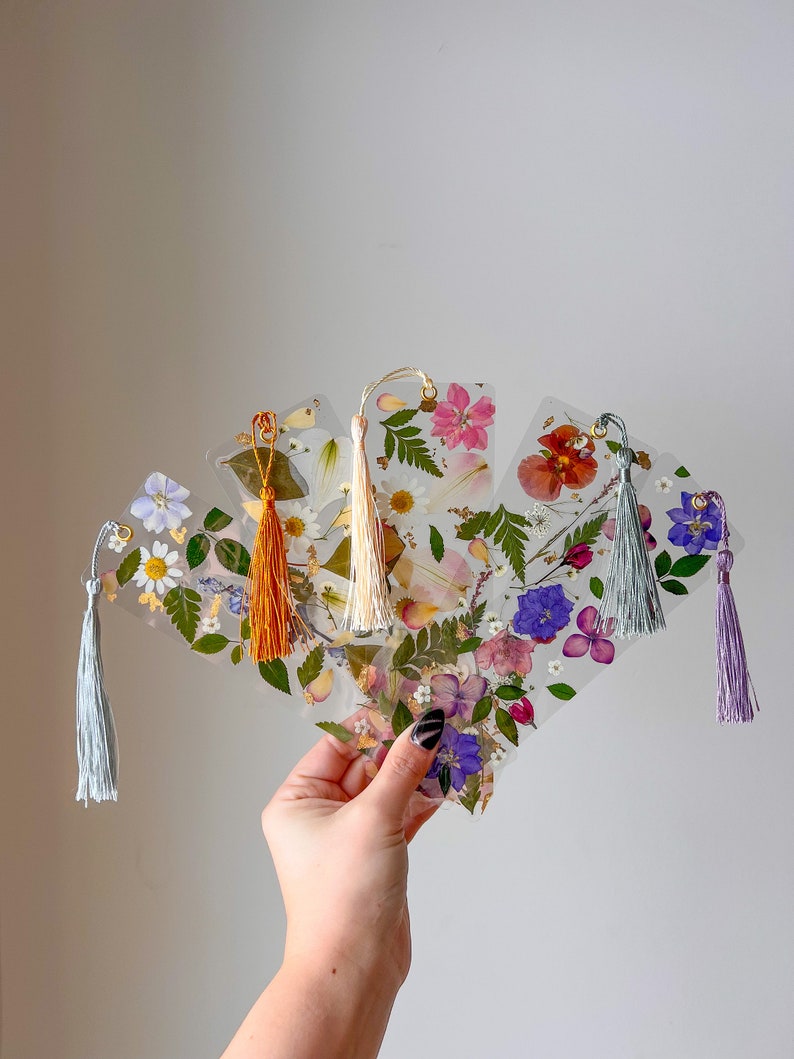 May include: Set of five pressed flower bookmarks with different floral designs and colors. Each bookmark has a tassel attached to the top.