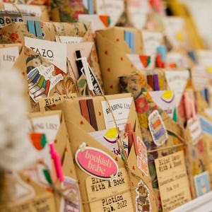 May include: A close-up of a shelf filled with wrapped gifts, each decorated with colorful stickers, twine, and paper. The gifts are labeled with titles like "Blind Date With A Book" and "Reading List".