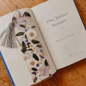 May include: A book titled "One Italian Summer" by Rebecca Serle with a pressed flower bookmark. The bookmark features a variety of dried flowers, including white daisies, purple flowers, and green leaves.