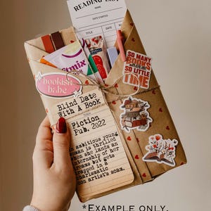 May include: A brown paper package wrapped with twine and decorated with hearts, stickers, and a book titled "Blind Date With A Book: Fiction Pub. 2022". The package also includes a reading list, a bookish babe sticker, and a sticker that says "So many books, so little time".
