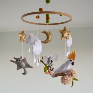 K&ouml;nnte beinhalten: Ein Holz-Babymobil mit Filztieren, darunter ein Koala, ein Gleitbeutler und ein Kakadu. Das Mobile enth&auml;lt auch Wolken, Sterne und einen Halbmond.