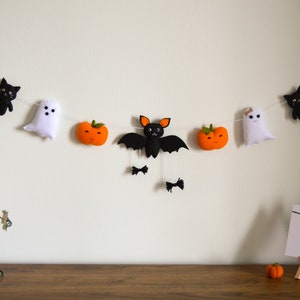 May include: A Halloween garland with felt decorations, including black cats, ghosts, pumpkins, and a bat. The garland is strung with white thread and orange pom-poms.