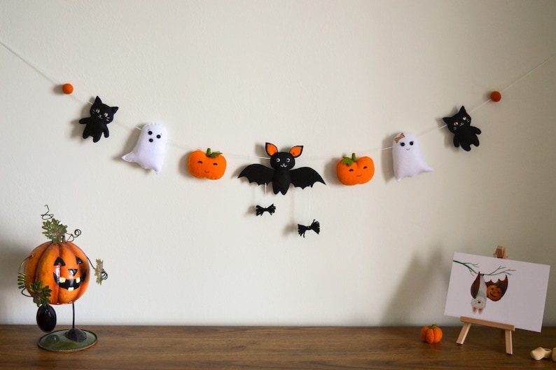 May include: A Halloween garland with felt decorations, including black cats, ghosts, pumpkins, and a bat. The garland is strung with white thread and orange pom-poms.