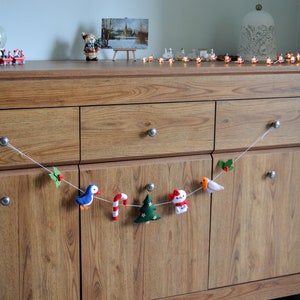 May include: A felt Christmas garland with a blue bird, candy cane, green Christmas tree, snowman, and a red bird. The garland is hanging on a wooden dresser with drawers.