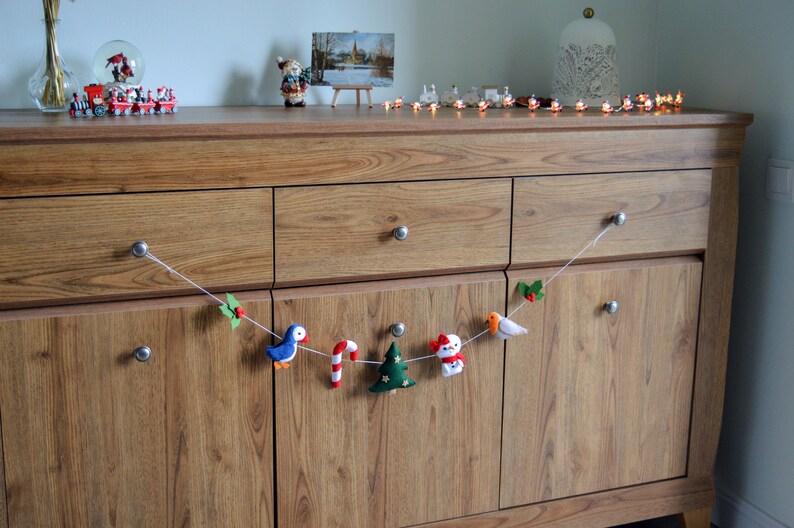 May include: A felt Christmas garland with a blue bird, candy cane, green Christmas tree, snowman, and a red bird. The garland is hanging on a wooden dresser with drawers.