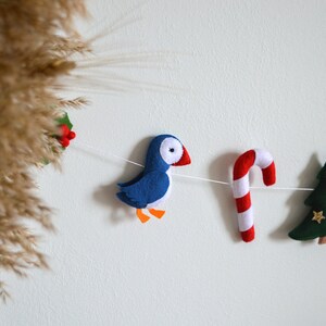 May include: A felt Christmas garland with a blue puffin, a red and white candy cane, a green Christmas tree, and a red and white holly sprig.