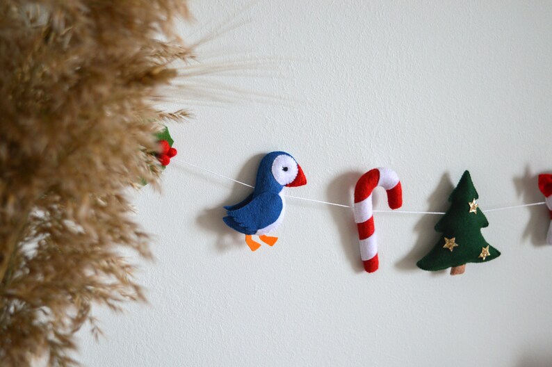May include: A felt Christmas garland with a blue puffin, a red and white candy cane, a green Christmas tree, and a red and white holly sprig.
