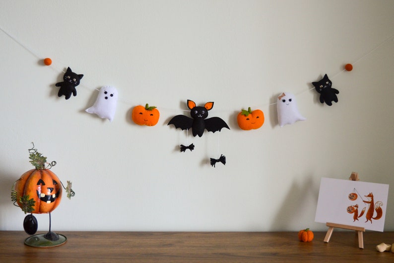 May include: A Halloween garland with black cats, ghosts, bats, and pumpkins. The garland is strung with white thread and has orange felt balls as spacers.