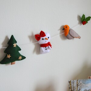 May include: A felt Christmas garland with a candy cane, a green Christmas tree with gold stars, a snowman with a red bow and scarf, a robin with red breast, and holly with red berries.