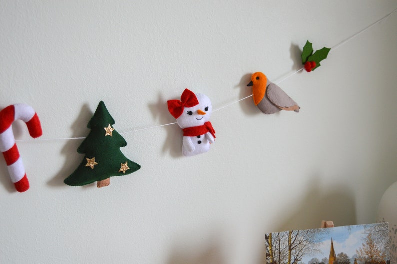 May include: A felt Christmas garland with a candy cane, a green Christmas tree with gold stars, a snowman with a red bow and scarf, a robin with red breast, and holly with red berries.