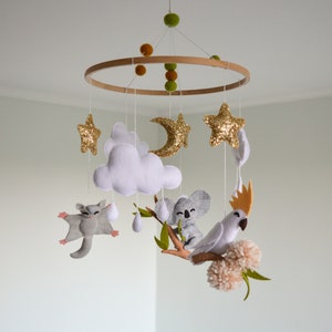 K&ouml;nnte beinhalten: Ein wei&szlig;es, graues und goldenes Baby-Mobile mit einem Holzring, Wolken, Sternen, einem Halbmond, einem Koala, einem Zuckergleiter und einem Kakadu.
