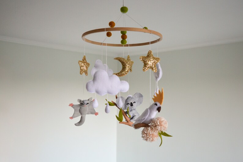 K&ouml;nnte beinhalten: Ein wei&szlig;es, graues und goldenes Baby-Mobile mit einem Holzring, Wolken, Sternen, einem Halbmond, einem Koala, einem Zuckergleiter und einem Kakadu.