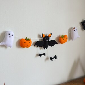 May include: A Halloween garland with felt decorations, including black cats, a ghost, a bat, and orange pumpkins. The garland is strung on a white string and is hanging on a white wall.