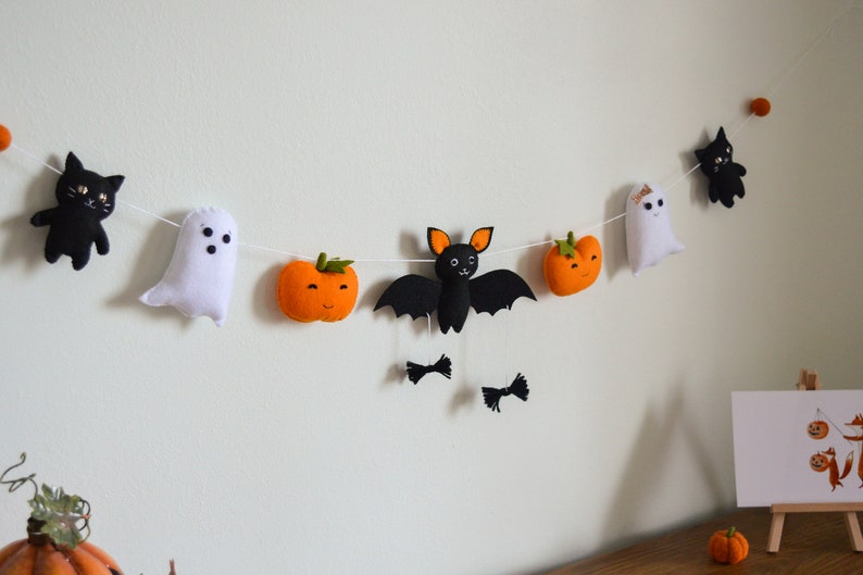 May include: A Halloween garland with felt decorations, including black cats, a ghost, a bat, and orange pumpkins. The garland is strung on a white string and is hanging on a white wall.