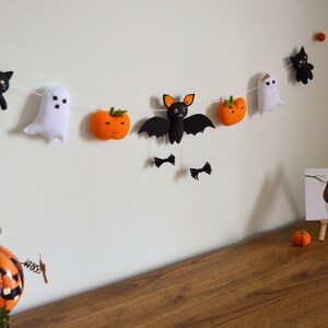 May include: A Halloween garland with felt decorations of a black cat, ghost, bat, and pumpkins. The garland is strung with white thread and is hanging on a white wall.