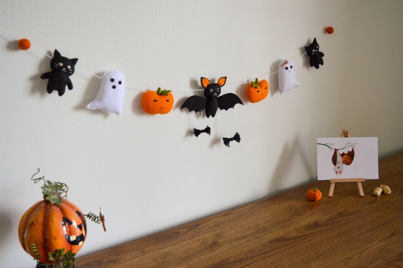 May include: A Halloween garland with felt decorations of a black cat, ghost, bat, and pumpkins. The garland is strung with white thread and is hanging on a white wall.