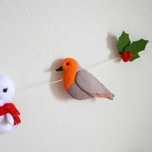 May include: A felt Christmas garland with a snowman, a robin, and holly. The snowman is white with a red bow and scarf. The robin is gray with an orange breast. The holly is green with red berries.
