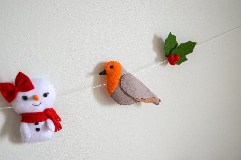 May include: A felt Christmas garland with a snowman, a robin, and holly. The snowman is white with a red bow and scarf. The robin is gray with an orange breast. The holly is green with red berries.