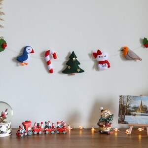 May include: A felt Christmas garland with a blue bird, candy cane, green Christmas tree, snowman, and a robin. The garland is hanging on a white wall.