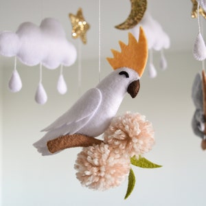 K&ouml;nnte beinhalten: Ein wei&szlig;er Filz-Babymobil mit einem wei&szlig;en Kakadu, einem grauen Koala, Wolken und Sternen. Der Kakadu sitzt auf einem braunen Ast mit zwei rosa Pompons.