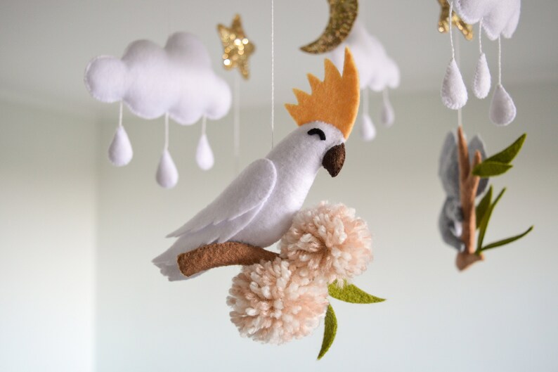 K&ouml;nnte beinhalten: Ein wei&szlig;er Filz-Babymobil mit einem wei&szlig;en Kakadu, einem grauen Koala, Wolken und Sternen. Der Kakadu sitzt auf einem braunen Ast mit zwei rosa Pompons.