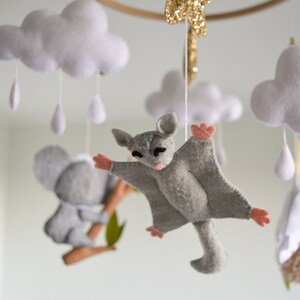 K&ouml;nnte beinhalten: Ein Baby-Mobile mit einem grauen Koala, einem grauen Gleitbeutler und wei&szlig;en Wolken mit Regentropfen. Das Mobile ist aus Filz gefertigt und hat einen Holzring.