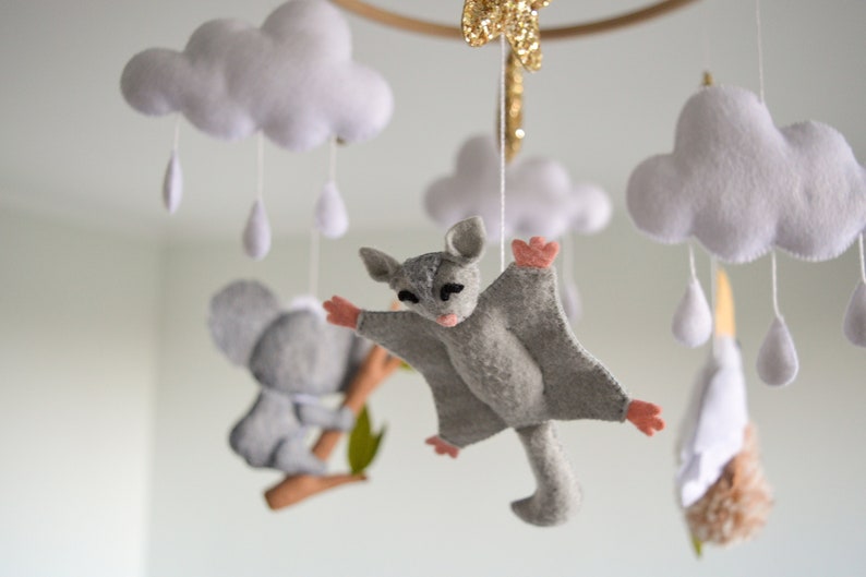 K&ouml;nnte beinhalten: Ein Baby-Mobile mit einem grauen Koala, einem grauen Gleitbeutler und wei&szlig;en Wolken mit Regentropfen. Das Mobile ist aus Filz gefertigt und hat einen Holzring.