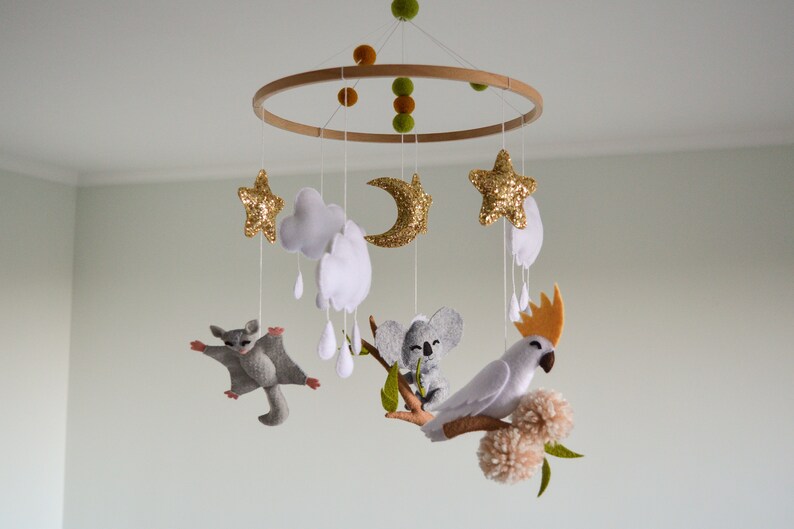 K&ouml;nnte beinhalten: Ein Holz-Babymobil mit Filztieren, darunter ein Koala, ein Gleitbeutler und ein Kakadu. Das Mobile enth&auml;lt auch Wolken, Sterne und einen Halbmond.