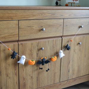 May include: A Halloween garland with felt bats, ghosts, and pumpkins hanging on a wooden dresser.