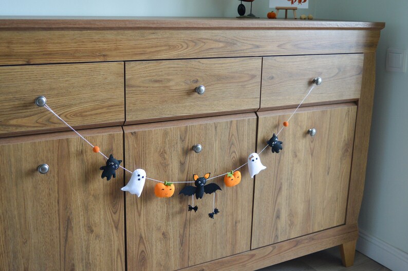 May include: A Halloween garland with felt bats, ghosts, and pumpkins hanging on a wooden dresser.