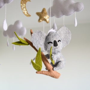 K&ouml;nnte beinhalten: Ein grauer Filz-Koala, der ein gr&uuml;nes Filzblatt h&auml;lt, h&auml;ngt an einem Ast mit gr&uuml;nen Filzbl&auml;ttern. Der Koala ist Teil eines Mobiles mit Wolken, Sternen und Regentropfen.