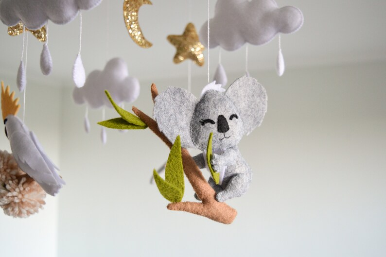 K&ouml;nnte beinhalten: Ein grauer Filz-Koala, der ein gr&uuml;nes Filzblatt h&auml;lt, h&auml;ngt an einem Ast mit gr&uuml;nen Filzbl&auml;ttern. Der Koala ist Teil eines Mobiles mit Wolken, Sternen und Regentropfen.