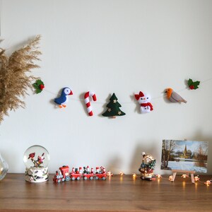 May include: A felt Christmas garland with a red and white candy cane, a green Christmas tree, a snowman, a blue bird, and a red robin. The garland is hanging on a white wall with a vase of dried pampas grass in the foreground.