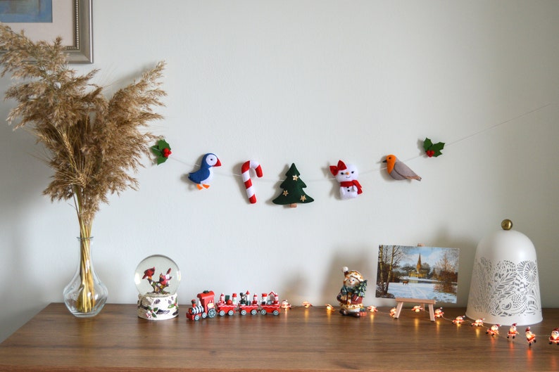 May include: A felt Christmas garland with a red and white candy cane, a green Christmas tree, a snowman, a blue bird, and a red robin. The garland is hanging on a white wall with a vase of dried pampas grass in the foreground.