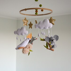 K&ouml;nnte beinhalten: Ein Holz-Babymobil mit einem wei&szlig;en Kakadu, einem Koala, Wolken, Sternen und Regentropfen. Das Mobile ist aus Filz gefertigt und hat einen naturbelassenen Holzring.