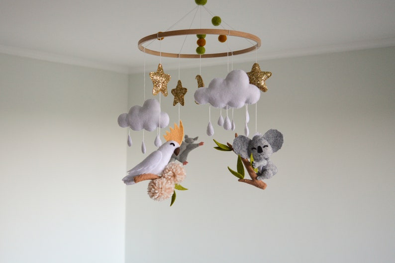 K&ouml;nnte beinhalten: Ein Holz-Babymobil mit einem wei&szlig;en Kakadu, einem Koala, Wolken, Sternen und Regentropfen. Das Mobile ist aus Filz gefertigt und hat einen naturbelassenen Holzring.